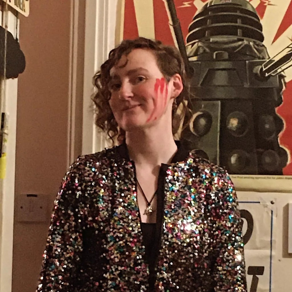 Jenny B posing in a sequined jacket in front of a Dalek poster, with red paint marks on side of face.
