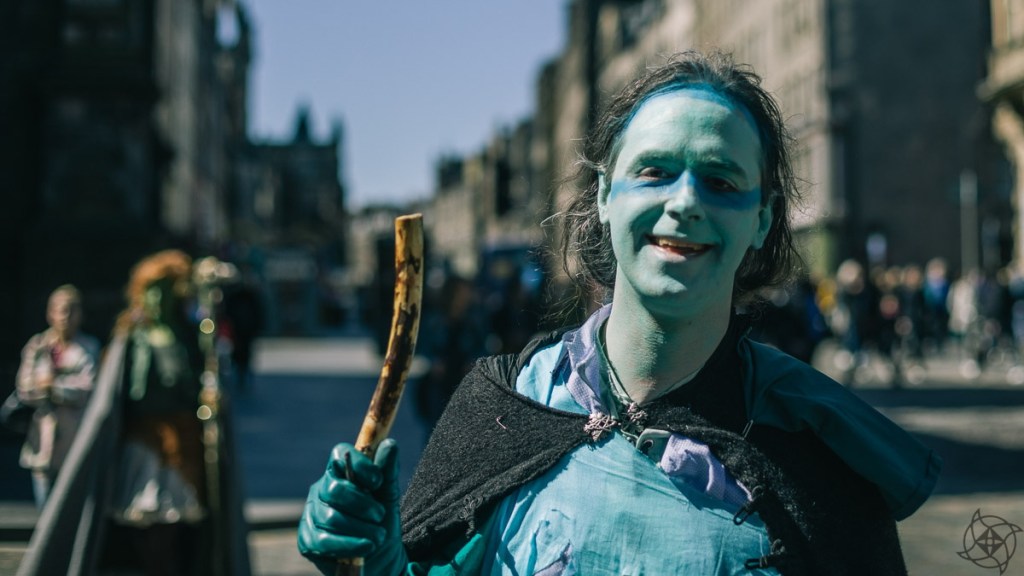 Robert wears a faerie costume, blue/green paint, and carries a wooden staff in teal gloves. In the background, people walk along a street of stone buildings.