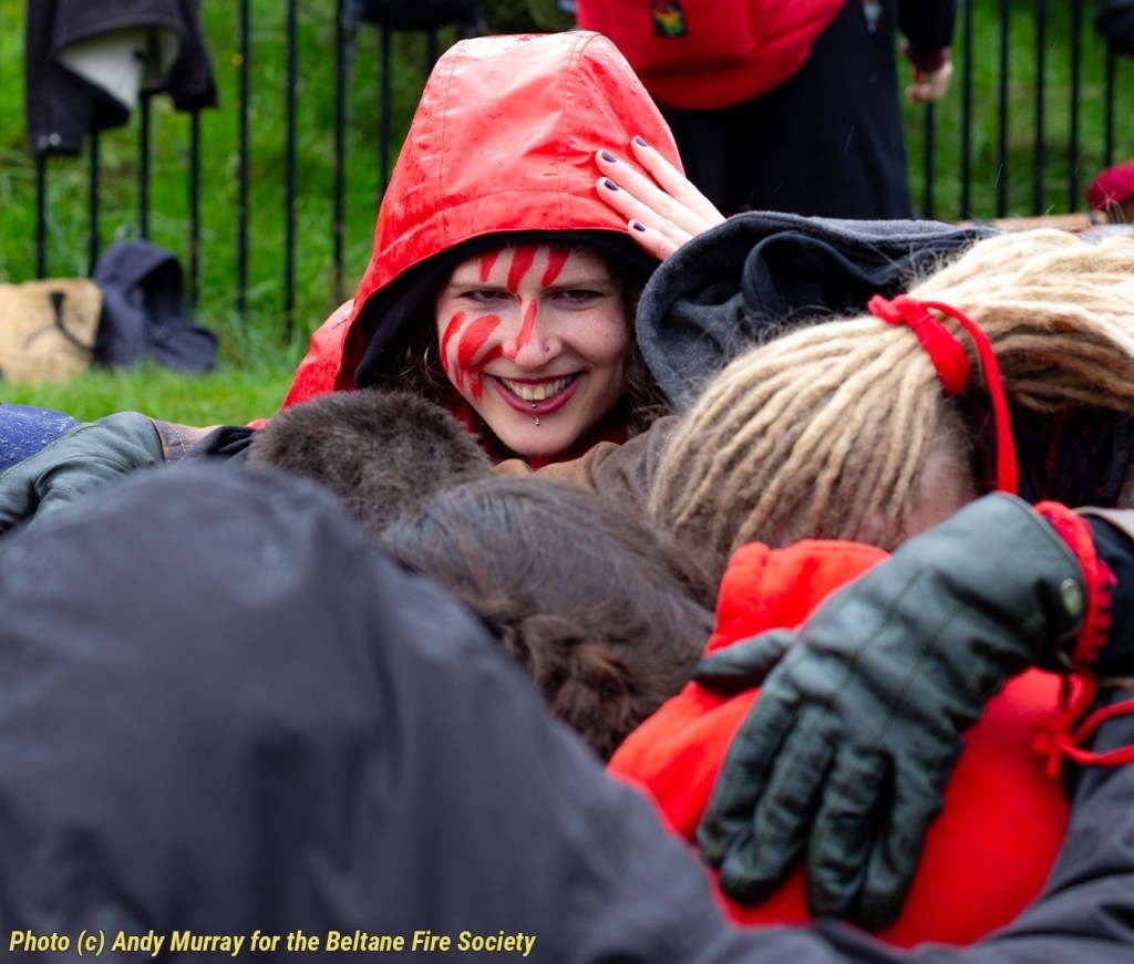 Outdoors. Steffie's head is seen in a red hood and red paint marks, emerging from a performers huddle. Grass and a black metal fence in the background.